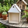 Domek dla owadów DIY ze sklejki 15 x 10 cm złożony na drewnianym stoliku w ogrodzie z roślinami w doniczkach w tle.