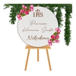 Personalizowana tablica powitalna na Komunię koło z białej płyty HDF 50 cm umieszczona na stojaku w otoczeniu dekoracji kwiatowych.