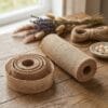 Wstążka Juta Dekoracyjna 25 cm x 5 m Naturalna Taśma Jutowa 180 g/m² - zdjęcie 2 Różne szerokości naturalnej wstążki jutowej 25 cm x 5 m obok sznurka i suszonych kwiatów na drewnianym stole.