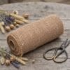 Wstążka z Juty Dekoracyjna 20 cm x 5 m Naturalna Ekologiczna 180 g/m² - zdjęcie 2 Gruba wstążka z juty dekoracyjna 20 cm x 5 m naturalna ekologiczna 180 g/m² na drewnianym stole obok nożyczek i suszonych kwiatów.